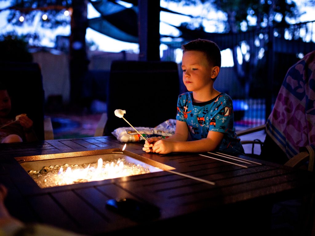A Boy Roasting A Marshmallow Over A Fire In His Backyard.